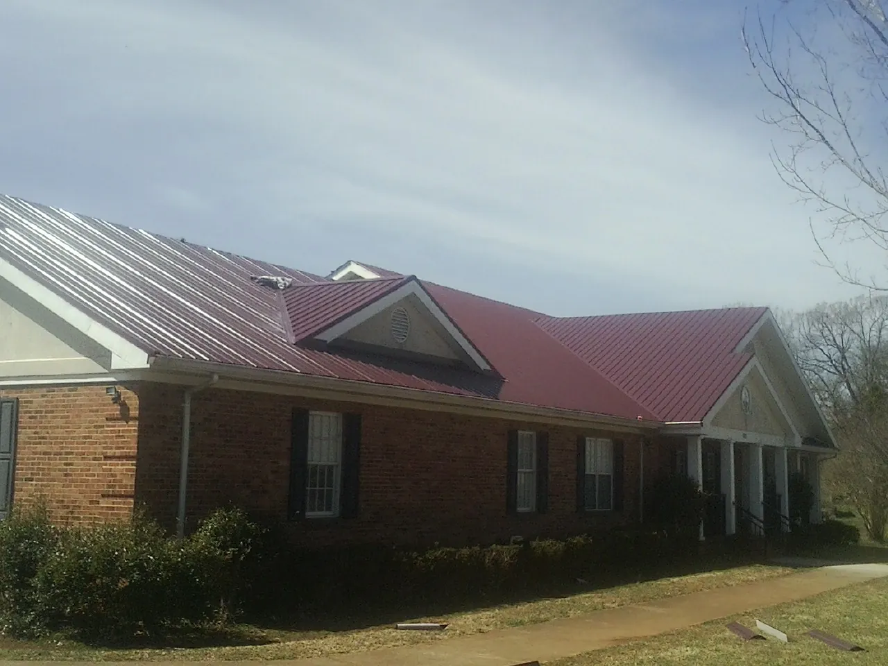 Red metal roof with gabled entries on a long brick building, from a Hail Damage Roof Repair job in Neotsu