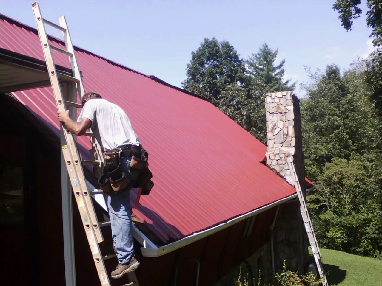 Roofer on a ladder beside a red metal roof and stone chimney — Soffit & Fascia Repair by Roofing Brothers in Neotsu