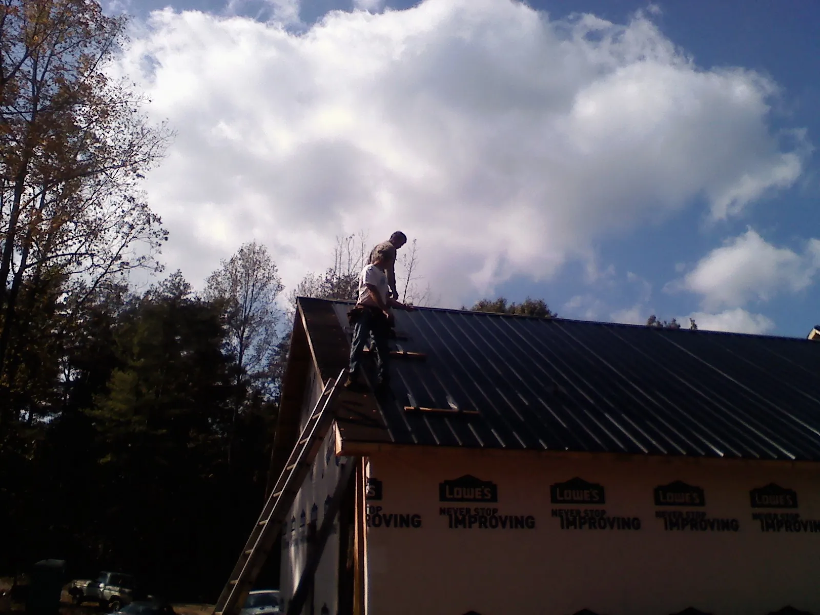 Roofer fastening dark metal panels high against a cloudy sky, from a Roof Inspection job in Neotsu